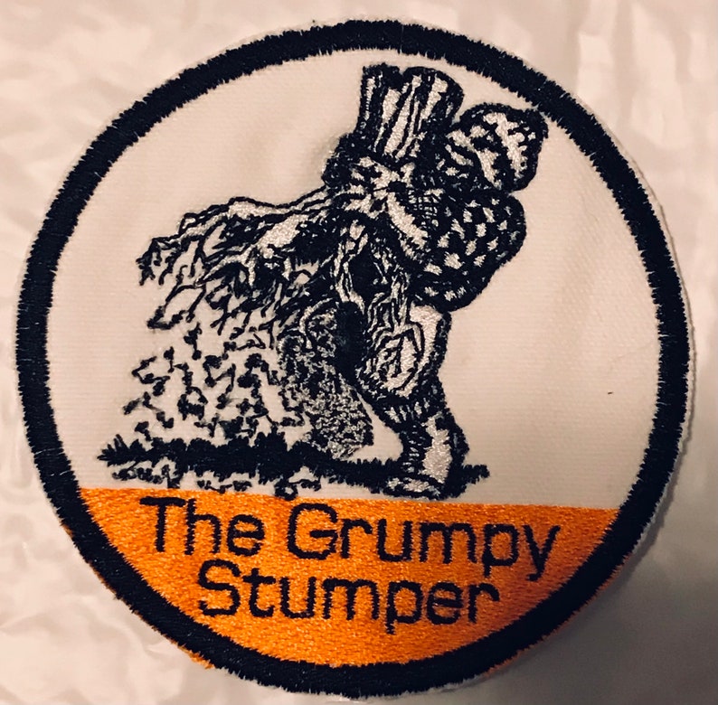K&ouml;nnte beinhalten: Ein wei&szlig;er runder Patch mit schwarzem Rand. Der Patch zeigt eine Schwarzwei&szlig;illustration einer m&uuml;rrisch aussehenden Figur mit einem gro&szlig;en Hut, die vor einem Baum steht. Der untere Teil des Patches hat ein orangefarbenes Band mit dem Text "The Grumpy Stumper" in Schwarz.