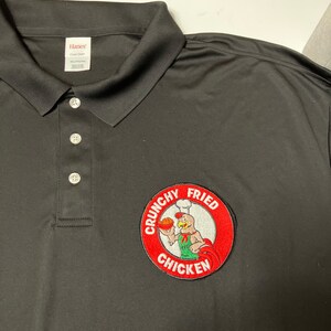 K&ouml;nnte beinhalten: Schwarzes Poloshirt mit einem roten, wei&szlig;en und gr&uuml;nen gestickten Aufn&auml;her mit der Aufschrift "Crunchy Fried Chicken" und einem Cartoon-Koch-Huhn, das ein Brath&auml;hnchenschenkel h&auml;lt.