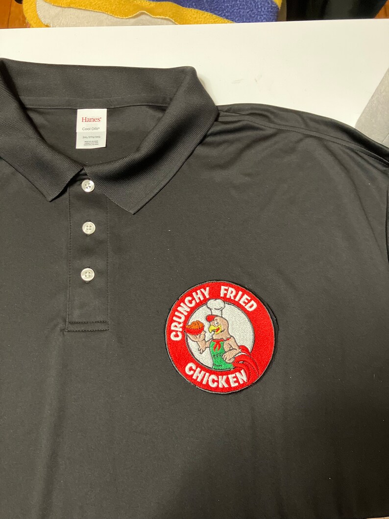 K&ouml;nnte beinhalten: Schwarzes Poloshirt mit einem roten, wei&szlig;en und gr&uuml;nen gestickten Aufn&auml;her mit der Aufschrift "Crunchy Fried Chicken" und einem Cartoon-Koch-Huhn, das ein Brath&auml;hnchenschenkel h&auml;lt.