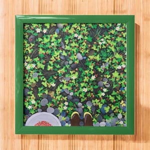 May include: A framed painting with a green border depicts a ground-level view of a garden. The scene includes green leaves, small white flowers, gray stones, and brown shoes. A small bowl of red berries is visible.