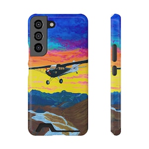 May include: A smartphone case featuring a painted scene of a small black airplane flying over a landscape of brown mountains and a white river. The sky is a vibrant gradient of yellow, orange, pink, and blue. The case has a black camera cutout.