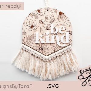 May include: A white wooden sign with a floral pattern and the words "be kind" in white lettering. The sign is attached to a white macrame tassel. The sign is laser cut and ready to be used.