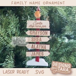 May include: A wooden Christmas ornament with the text "Family Name Ornament." The ornament features a signpost design with the words "Santa stop here for" and personalized names. The ornament is decorated with a red and white color scheme and a cloud base.