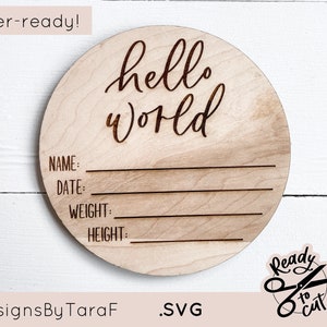 May include: A laser-cut wooden circle with the text "hello world" in cursive script. The circle has lines for writing the baby's name, date, weight, and height. The text "Designs By TaraF" and ".SVG" are at the bottom of the circle. A small circle with a pair of scissors and the text "Ready to Cut" is in the lower right corner.