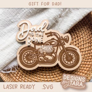 Puede incluir: Un letrero de madera cortado con láser con el texto "Dad to the Bone" y un diseño de motocicleta. El letrero está en una cesta tejida con un paño blanco.