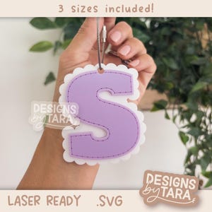 Puede incluir: Adorno con forma de letra "S" de color lavanda, con borde festoneado y detalles cosidos, suspendido de un cordón plateado. El texto en la imagen dice "3 tamaños incluidos!" y "LASER READY .SVG". De "DESIGNS by TARA".