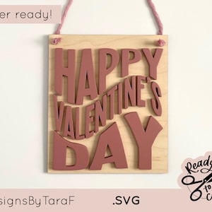 May include: A wooden sign with the words "Happy Valentine's Day" in pink lettering. The sign is laser cut and ready to be assembled. The sign is hanging from a pink string.