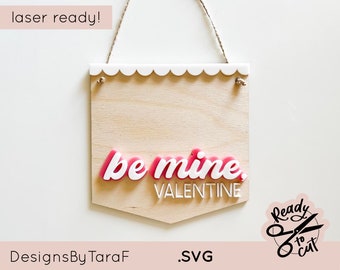 May include: A wooden sign with a scalloped white border and the text "be mine, VALENTINE" in pink and white letters. The sign is laser cut and ready to be assembled. The image also includes a logo with the text "Ready to cut" and a pair of scissors.