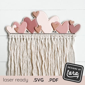 Könnte beinhalten: Wanddekoration aus Holz mit geschichteten Herzen in Rosa- und Beigetönen. Darunter befindet sich ein Makramee-Fransen. Der Text "laser ready .SVG .PDF" befindet sich unten links. Das Logo "DESIGNS BY tara" befindet sich unten rechts.