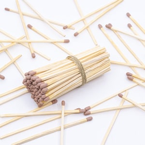 May include: A bundle of wooden matches with brown tips, tied together with twine, surrounded by loose matches on a white surface.