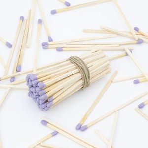 May include: A bundle of wooden matches with purple tips, tied together with twine, surrounded by loose matches on a white surface.