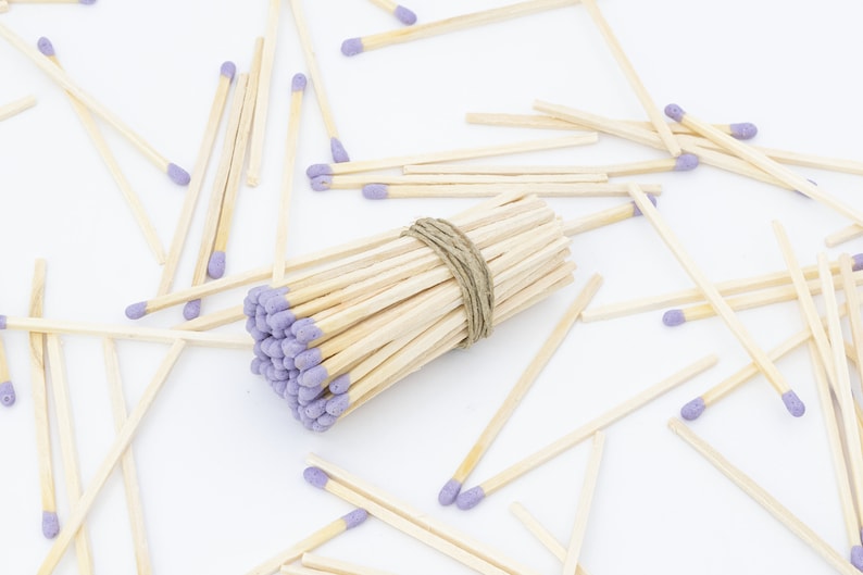 May include: A bundle of wooden matches with purple tips, tied together with twine, surrounded by loose matches on a white surface.