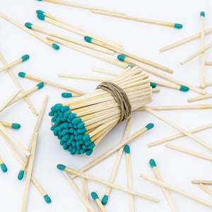 May include: A bundle of wooden matches with teal colored tips, tied together with twine. The matches are scattered around the bundle on a white surface.