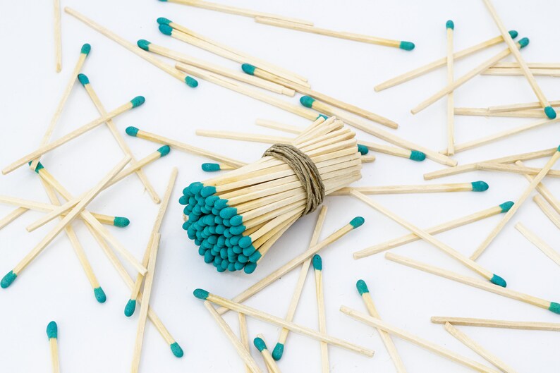 May include: A bundle of wooden matches with teal colored tips, tied together with twine. The matches are scattered around the bundle on a white surface.