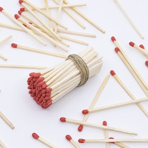 May include: A bundle of red-tipped wooden matches tied together with twine, surrounded by loose matches on a white surface.