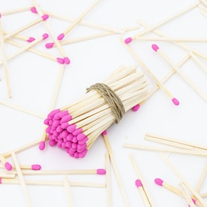 May include: A bundle of wooden matches with pink tips, tied together with twine, surrounded by loose matches on a white surface.