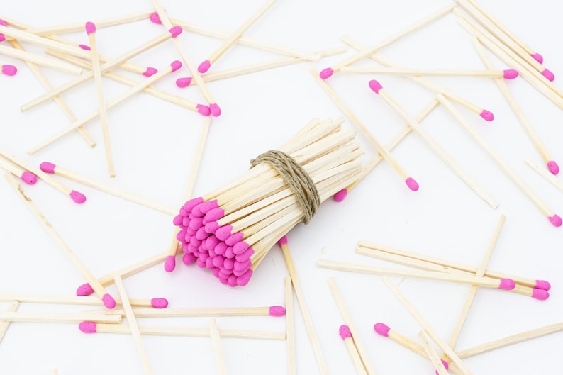 May include: A bundle of wooden matches with pink tips, tied together with twine, surrounded by loose matches on a white surface.