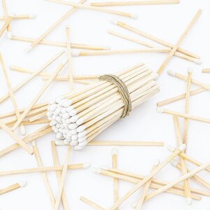May include: A bundle of white tipped wooden matches tied together with twine, surrounded by loose matches on a white surface.