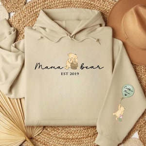 K&ouml;nnte beinhalten: Ein beiger Hoodie mit einer Winnie Puuh-Figur, die einen Honigtopf h&auml;lt, und dem Text "Mama Bear EST 2019". Der rechte &Auml;rmel zeigt eine Winnie Puuh-Figur, die einen Ballon mit der Aufschrift "Callie Mae" h&auml;lt.