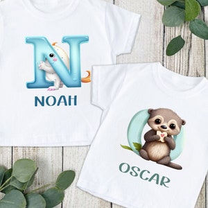 Peut inclure: Deux t-shirts blancs avec des lettres bleues et vertes et des illustrations d'animaux. Le premier t-shirt a une lettre N bleue avec un monstre blanc et le nom Noah. Le deuxième t-shirt a une lettre O verte avec une loutre brune et le nom Oscar.