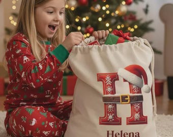 Puede incluir: Una niña en pijama rojo navideño abre un saco de Papá Noel personalizado. El saco es beige con un diseño de letra "H" roja, un gorro de Papá Noel y el nombre "Helena" impreso. Un árbol de Navidad decorado está en el fondo.