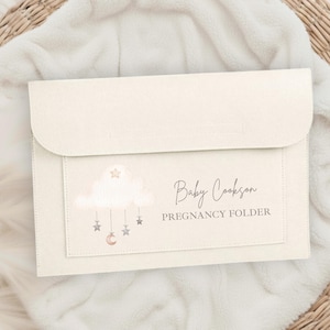 Può includere: Un raccoglitore per la gravidanza color crema con chiusura a patta. La parte anteriore presenta un disegno di nuvole con stelle sospese e una luna crescente. Il testo "Baby Cookson PREGNANCY FOLDER" è stampato.