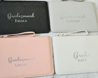 May include: Four different clutches with zipper closures and wrist straps. The clutches are in black, grey, pink, and white. The clutches are personalised with the words "Bride" and "Bridesmaid" with names in silver lettering.