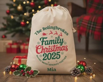 May include: A cream-colored drawstring Christmas gift bag with the text "The Bolingbrokes Family Christmas 2025" in green and red. The bag also has the name "Mia" printed on it. The bag is surrounded by Christmas decorations.