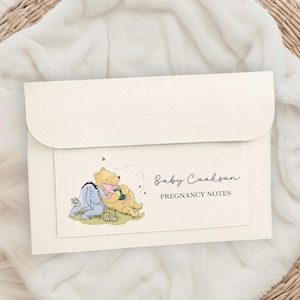Può includere: Porta note di gravidanza color crema con chiusura a patta. La parte anteriore presenta un'illustrazione di Winnie the Pooh e il testo "Baby Cookson PREGNANCY NOTES". Il supporto è su una superficie bianca strutturata.