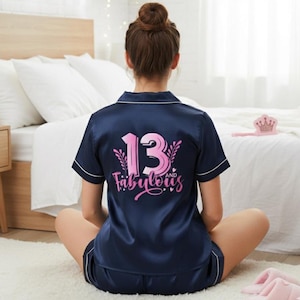 May include: Navy blue satin pyjama set with white trim. The top displays the text "13 and Fabulous" in pink. The person is sitting on a white rug in front of a bed with white bedding. Pink slippers are visible.