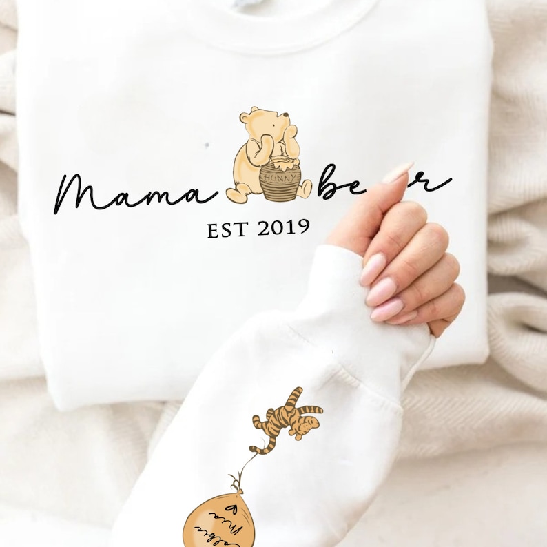 May include: White sweatshirt with a Winnie the Pooh inspired design. The text "Mama bear EST 2019" is printed on the front of the sweatshirt. A cartoon image of Winnie the Pooh holding a honey pot is above the text. A cartoon image of Tigger holding onto a balloon is on the sleeve of the sweatshirt.