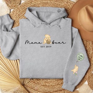 K&ouml;nnte beinhalten: Grauer Kapuzenpullover mit einem Winnie-the-Pooh-inspirierten Design. Die Vorderseite des Pullovers zeigt einen B&auml;ren, der einen Honigtopf h&auml;lt, und den Text "Mama bear EST 2019". Der linke &Auml;rmel zeigt einen B&auml;ren, der einen Ballon mit dem Text "Cubs  Me" h&auml;lt.