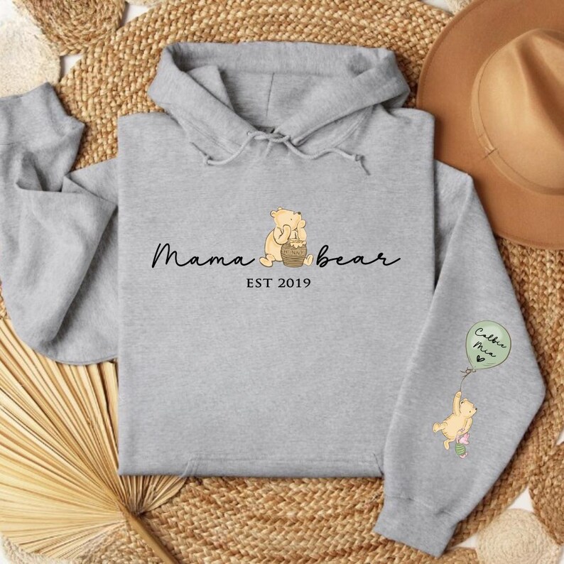 K&ouml;nnte beinhalten: Grauer Kapuzenpullover mit einem Winnie-the-Pooh-inspirierten Design. Die Vorderseite des Pullovers zeigt einen B&auml;ren, der einen Honigtopf h&auml;lt, und den Text "Mama bear EST 2019". Der linke &Auml;rmel zeigt einen B&auml;ren, der einen Ballon mit dem Text "Cubs  Me" h&auml;lt.