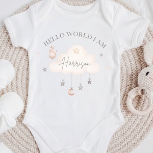 Puede incluir: Body de bebé blanco con el texto "HELLO WORLD I AM" sobre una ilustración de nube con el nombre "Harrison". La nube tiene estrellas colgantes y una luna creciente. También aparece un pequeño cerdo con un globo.