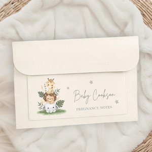 Può includere: Cartella per note di gravidanza color crema con chiusura a patta. La parte anteriore presenta una tasca con un'illustrazione di animali impilati: giraffa, leone e agnello. Il testo "Baby Cookson" e "Pregnancy Notes" è stampato.