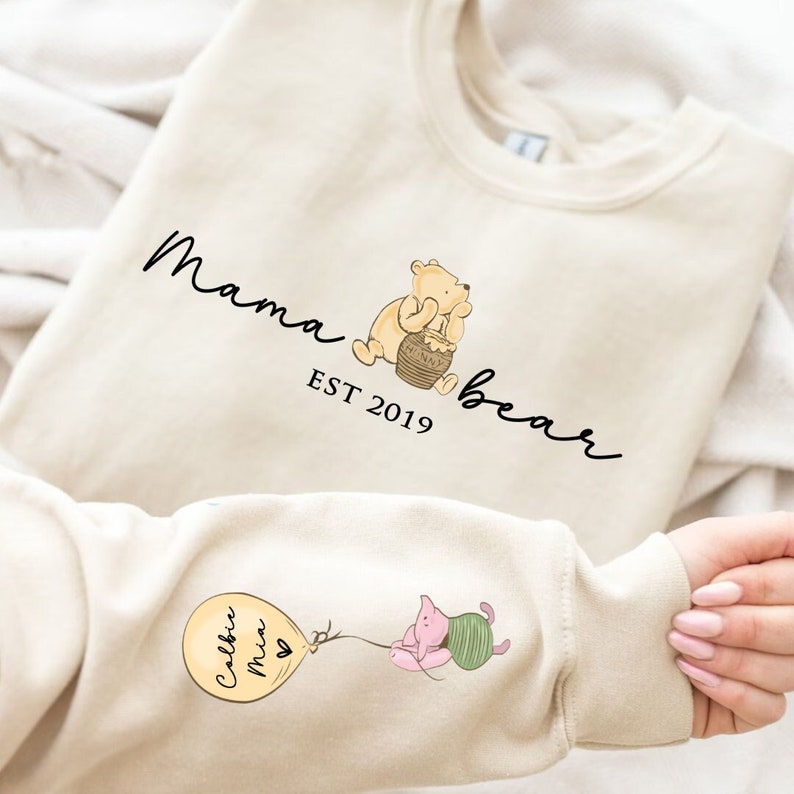 May include: A cream-colored sweatshirt with the text "Mama bear est 2019" and an illustration of Winnie the Pooh holding a honey pot. The sweatshirt also features a small illustration of Piglet holding a balloon with the text "Callie Mia" on it.