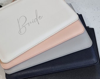 May include: Four clutch bags in different colours, stacked on top of each other. The top bag is white with the word "Bride" written in silver script. The other bags are pink, grey, and black.