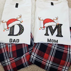 Puede incluir: Dos camisetas blancas con pantalones de pijama a cuadros a juego. Las camisetas presentan un diseño temático navideño con un gorro de Papá Noel rojo, cuernos y las letras "D" y "M" con las palabras "DAD" y "MOM" debajo. Los pantalones de pijama son a cuadros rojos, negros y blancos.