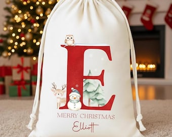 Puede incluir: Una bolsa de regalo navideña de color crema con la letra «E» roja e ilustraciones festivas. La bolsa presenta un muñeco de nieve, un ciervo, un búho y un árbol de Navidad. El texto «FELIZ NAVIDAD» y un nombre están impresos debajo.