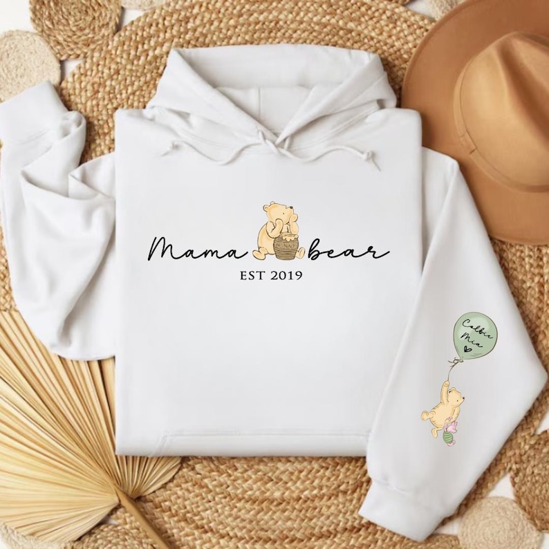 K&ouml;nnte beinhalten: Wei&szlig;er Hoodie mit einem Winnie-the-Pooh-inspirierten Design. Der Text lautet "Mama Bear Est 2019". Ein kleiner Winnie-the-Pooh-Charakter h&auml;lt einen Honigtopf. Der linke &Auml;rmel hat einen kleinen Winnie-the-Pooh-Charakter, der einen Ballon h&auml;lt, auf dem "Cubs Me" steht.