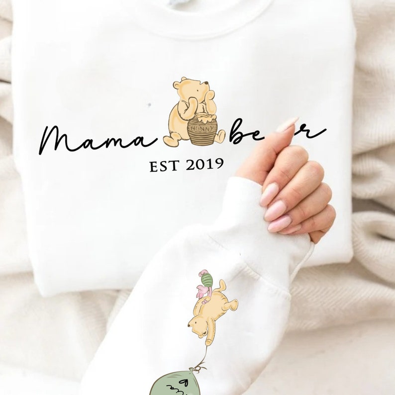 May include: White sweatshirt with a Winnie the Pooh inspired design. The text "Mama bear EST 2019" is printed on the front of the sweatshirt. The design features a cartoon Winnie the Pooh holding a honey pot and another Winnie the Pooh holding a balloon.