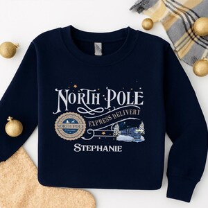 May include: Navy blue sweatshirt featuring a North Pole Express Delivery design. The design includes the words "North Pole", a train illustration, and the name "Stephanie". The sweatshirt has a crew neck and long sleeves.
