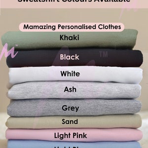 May include: A stack of sweatshirts in various colors, including khaki, black, white, ash, grey, sand, light pink, and light blue. The text "Sweatshirt Colours Available" is at the top of the image, and the text "Mamazing Personalised Clothes" is in the middle of the image.