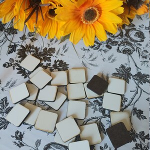 May include: A collection of 20 white square wooden tiles with black edges, scattered on a black and white floral patterned surface. The tiles are blank and ready for crafting or painting.