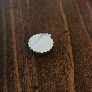 Pu&ograve; includere: Un adesivo rotondo di colore argento con un bordo bianco a forma di conchiglia. L'adesivo &egrave; parzialmente staccato, rivelando un retro adesivo bianco.