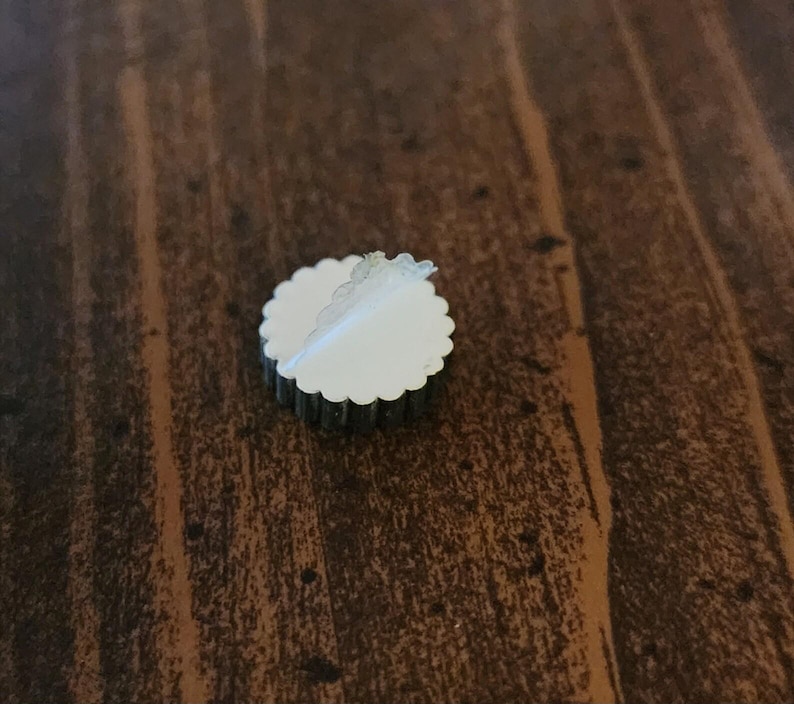 Pu&ograve; includere: Un adesivo rotondo di colore argento con un bordo bianco a forma di conchiglia. L'adesivo &egrave; parzialmente staccato, rivelando un retro adesivo bianco.