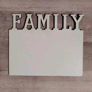 Könnte beinhalten: Ein Holzschild mit dem Wort "FAMILY" in weißen Buchstaben auf beigefarbenem Hintergrund. Das Schild hat einen leeren Raum unter dem Wort, um Fotos oder andere Dekorationen hinzuzufügen.