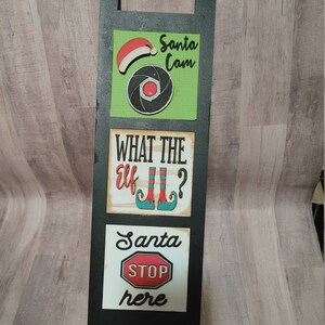 May include: A black wooden stand with three signs. The top sign is green with a red Santa hat and a camera lens. The middle sign is white with the text "What the elf Jill?" and a pair of elf shoes. The bottom sign is white with the text "Santa Stop here" and a red stop sign.