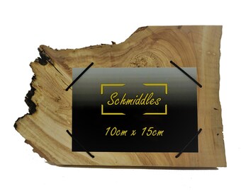Könnte beinhalten: Ein Rahmen aus Naturholz mit Baumkante und einem rechteckigen schwarzen Mittelteil mit gelbem Text "Schmiddles" und "10cm x 15cm". Das Holz hat eine hellbraune Maserung mit dunklen, unregelmäßigen Rindenkanten auf der linken Seite und vier schwarzen Eckclips.