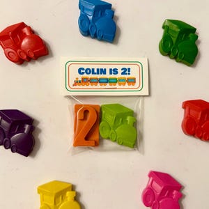 Peut inclure: Des aimants en forme de train jouet colorés en rouge, bleu, vert, violet, jaune et rose. Un sac en plastique transparent contient le chiffre 2 orange et un train vert. Une carte indique "COLIN IS 2!"